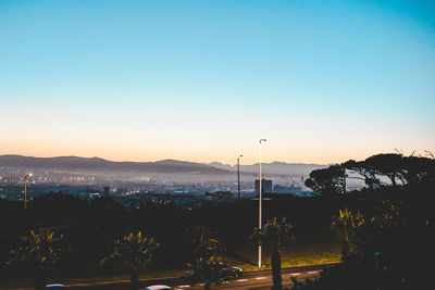 Illuminated street by silhouette mountains against clear sky at sunset