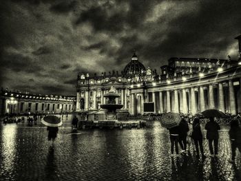 Illuminated buildings by river against cloudy sky