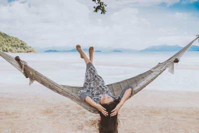 Woman liying in hammock with arms raised on beach against sky