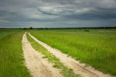 Sandy road and green fields, dark clouds on the sky, spring rural view