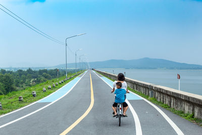 Rear view of mother and son riding bicycle on road against sky