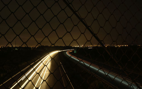 Light trails on highway seen through chainlink fence against sky at night