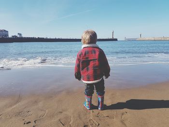 Rear view of boy standing on sea shore against clear blue sky