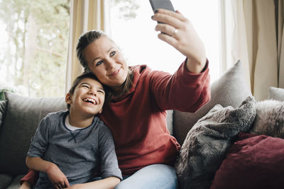 Mother taking selfie with happy autistic son while sitting on sofa at home