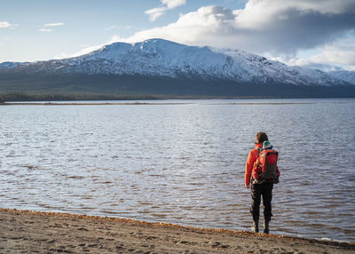 Hiker carrying child in backpack standing at the shore facing a snow capped mountain