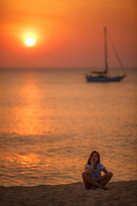 Girl sitting in sea against orange sunset sky and  yacht