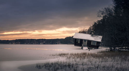 Cottage on frozen landscape against sky during sunset