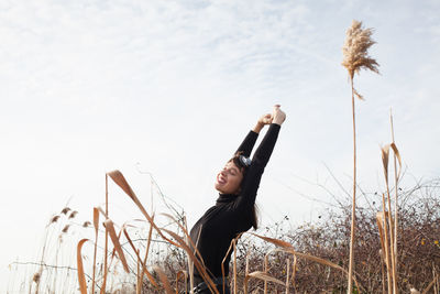 Smiling young woman standing with arms raised on field against sky