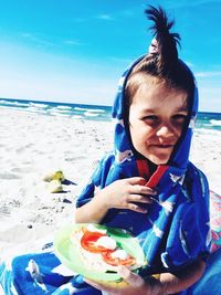 Portrait of cute boy eating food while sitting at beach against blue sky during sunny day