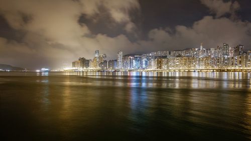 Illuminated city at waterfront against cloudy sky