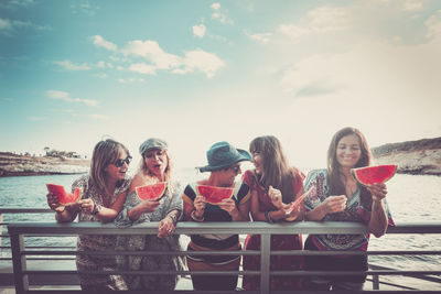 Women eating watermelon slice while standing by railing against sky