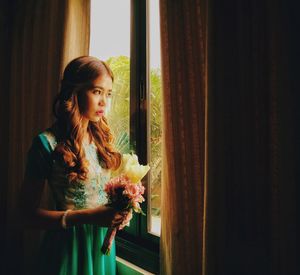 Bride holding bouquet looking through window while standing at home