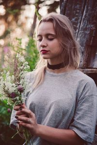 Beautiful young woman holding flowers while standing against tree