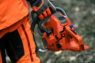 Close up view of an hand holding a chainsaw. lumberjack at work wears orange protective equipment. 