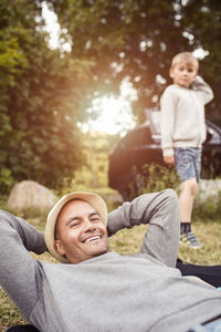 Portrait of happy man relaxing with boy standing at background in park