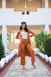 Full body of barefoot young ethnic female model wearing stylish brown suit looking at camera while standing on stone pathway against oriental building