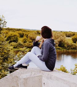 Side view of woman sitting on retaining wall while looking at lake against sky