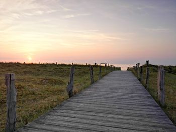 Wooden boardwalk on landscape against sky during sunset