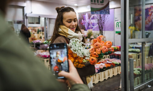 A woman takes a photo with her mobile phone of another woman with a bunch of peonies