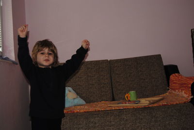 Portrait of cute boy with arms raised standing by sofa at home