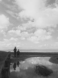 Friends standing by reflection in water at beach against cloudy sky