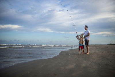 Playful father and sons with fishing rod standing at beach against cloudy sky