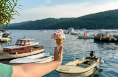 Personal perspective of hand holding two ice cream in cone on waterfront with moored boats in sea