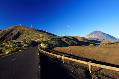 Road leading towards green mountain against clear blue sky