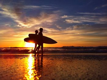 Silhouette couple carrying surfboards on beach at sunset