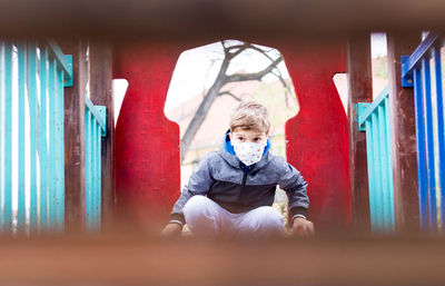 Small boy wearing protective face mask while playing on the playground during coronavirus pandemic.