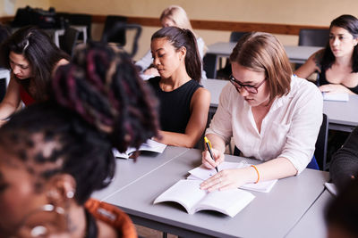 High angle view of female student writing in book while sitting with classmates in classroom