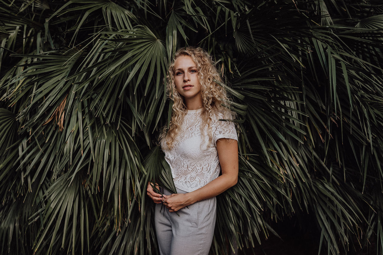 one person, young adult, standing, real people, front view, plant, growth, leisure activity, tree, portrait, looking at camera, leaf, nature, lifestyles, casual clothing, palm tree, waist up, young women, outdoors, hairstyle, contemplation, palm leaf, beautiful woman