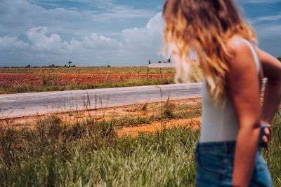 Side view of female traveler in casual wear standing on crossroad with brown soil among green tropical plants under grey cloudy heaven in cuba