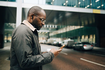 Side view of businessman using mobile phone while standing on street against building in city