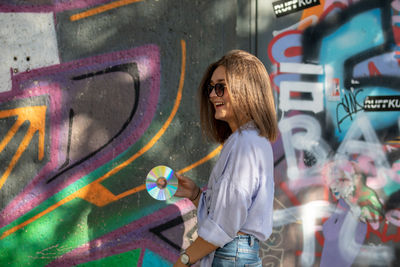 Beautiful woman holding compact disc while standing against graffiti wall