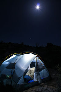 Man resting in illuminated tent against sky at night