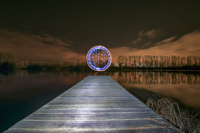 Illuminated ferris wheel by lake against sky at night