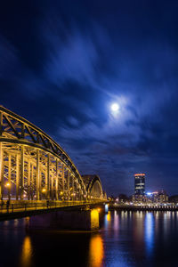 Illuminated bridge over river by buildings against sky at night