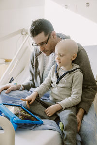 Father recovering from surgery watches tablet with son on hospital bed