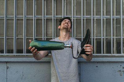 Young man smiling and demonstrating leg prosthesis while standing near building on city street