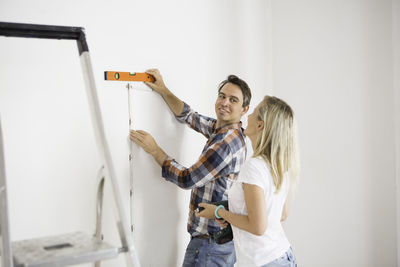 Man looking at girlfriend while measuring white wall at home