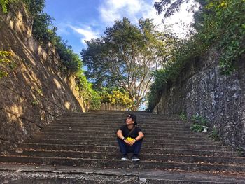 Young man sitting on staircase by trees against sky