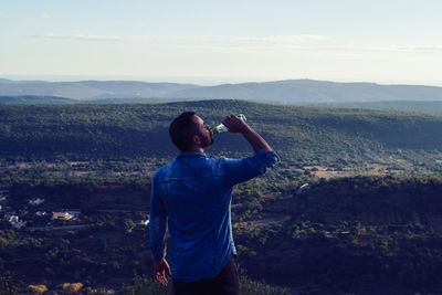 Man drinking from bottle while standing on landscape against sky