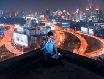 Businessman talking on mobile phone while using laptop at building terrace against illuminated city