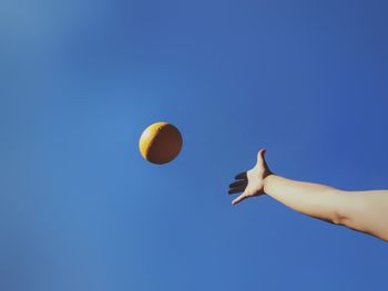 Low angle view of a hand throwing a yellow ball against blue sky