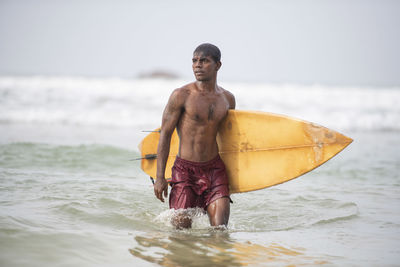 Shirtless man with surfboard wading in sea against sky