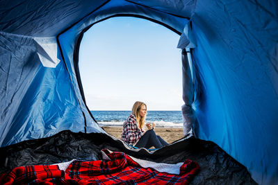 Young woman sitting outside tent by sea