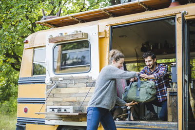 Smiling woman assisting man in unloading luggage from yellow motor home during camping in forest