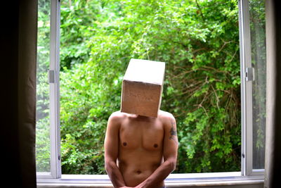 Shirtless man wearing box while standing against window
