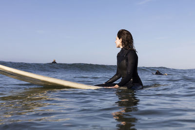 Thoughtful female surfer sitting on surfboard in sea against clear sky